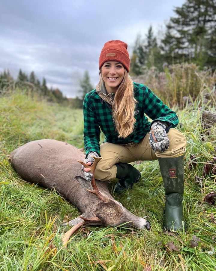 Woman kneels next to a harvested deer in a field, wearing hunting gear; cloudy day.