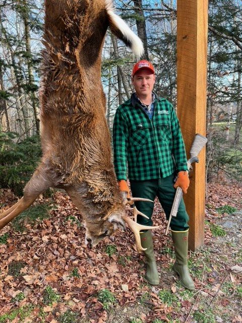 Man in green plaid shirt stands with a deer carcass hanging from a beam; he holds a rifle, wearing a red hat.