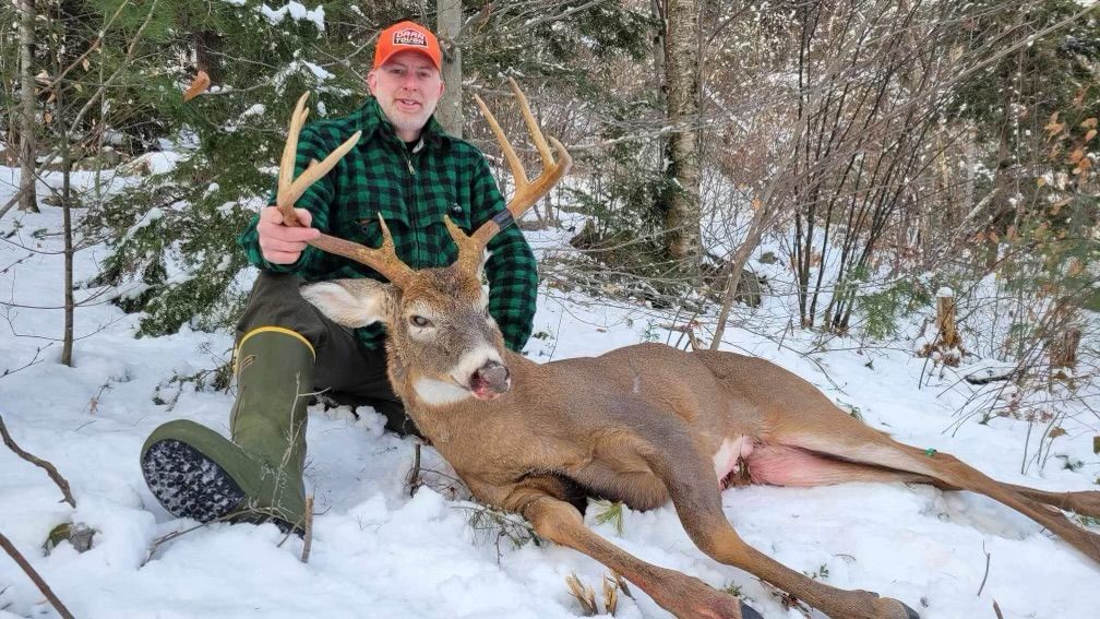 Man in orange hat and green plaid shirt sits next to a large deer with antlers in snowy woods.