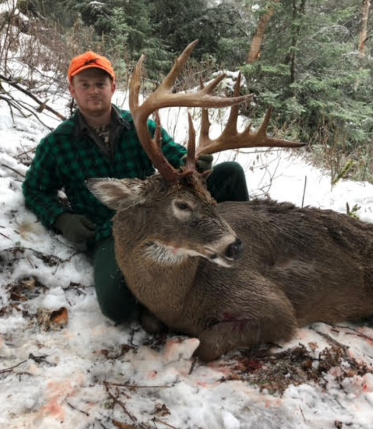 Hunter kneels next to a large deer with impressive antlers, both on snow. Hunter wears orange cap, plaid shirt.