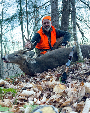 Hunter kneels beside a dead deer in forest. He wears orange safety gear and holds deer antlers.