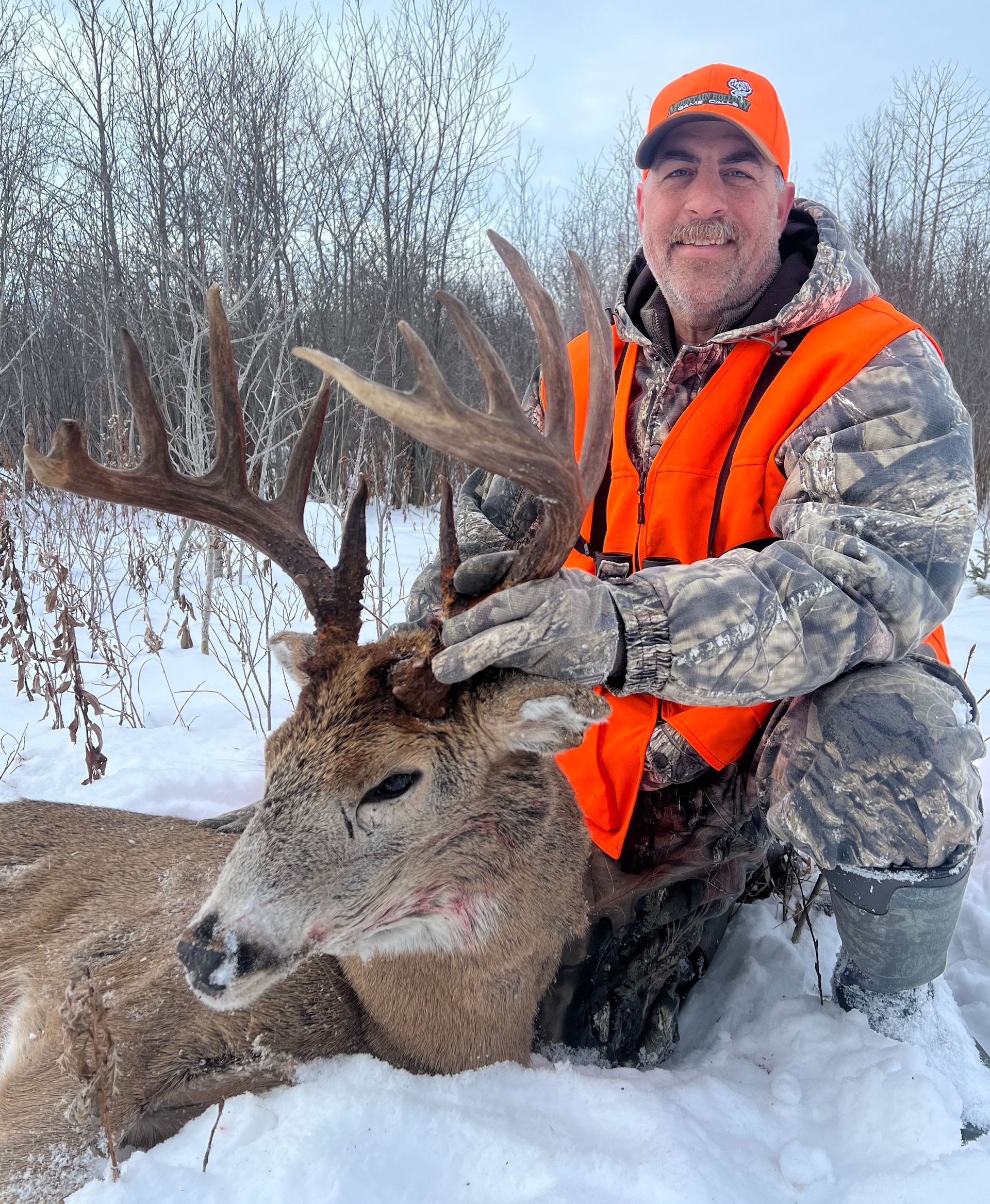 Hunter kneels beside a large buck with impressive antlers, both on snow. Man wears orange vest and hat.