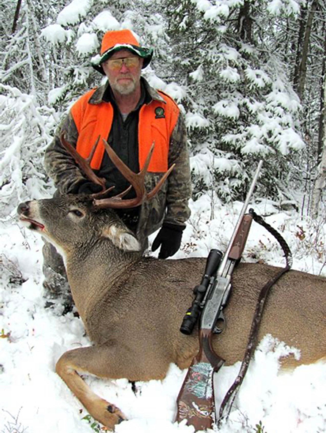 Hunter in orange vest with rifle poses with a deer in snowy woods.
