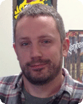 Man with short hair and a beard, wearing a plaid shirt, smiling in a room with posters.