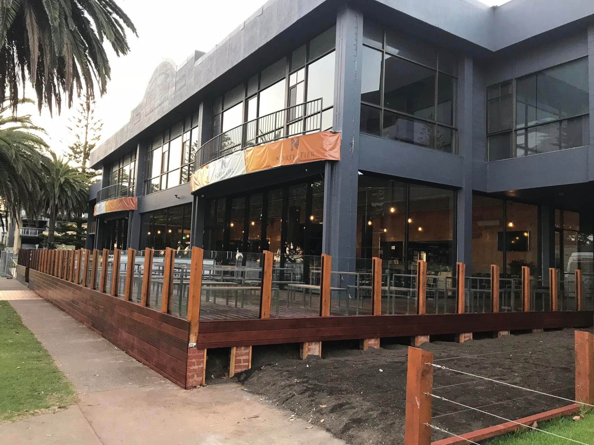 Modern two-story building with glass storefronts and a wooden deck along a tree-lined sidewalk
