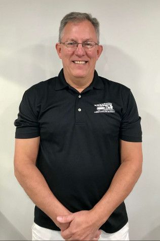 Smiling man in a black polo shirt standing against a plain light wall.