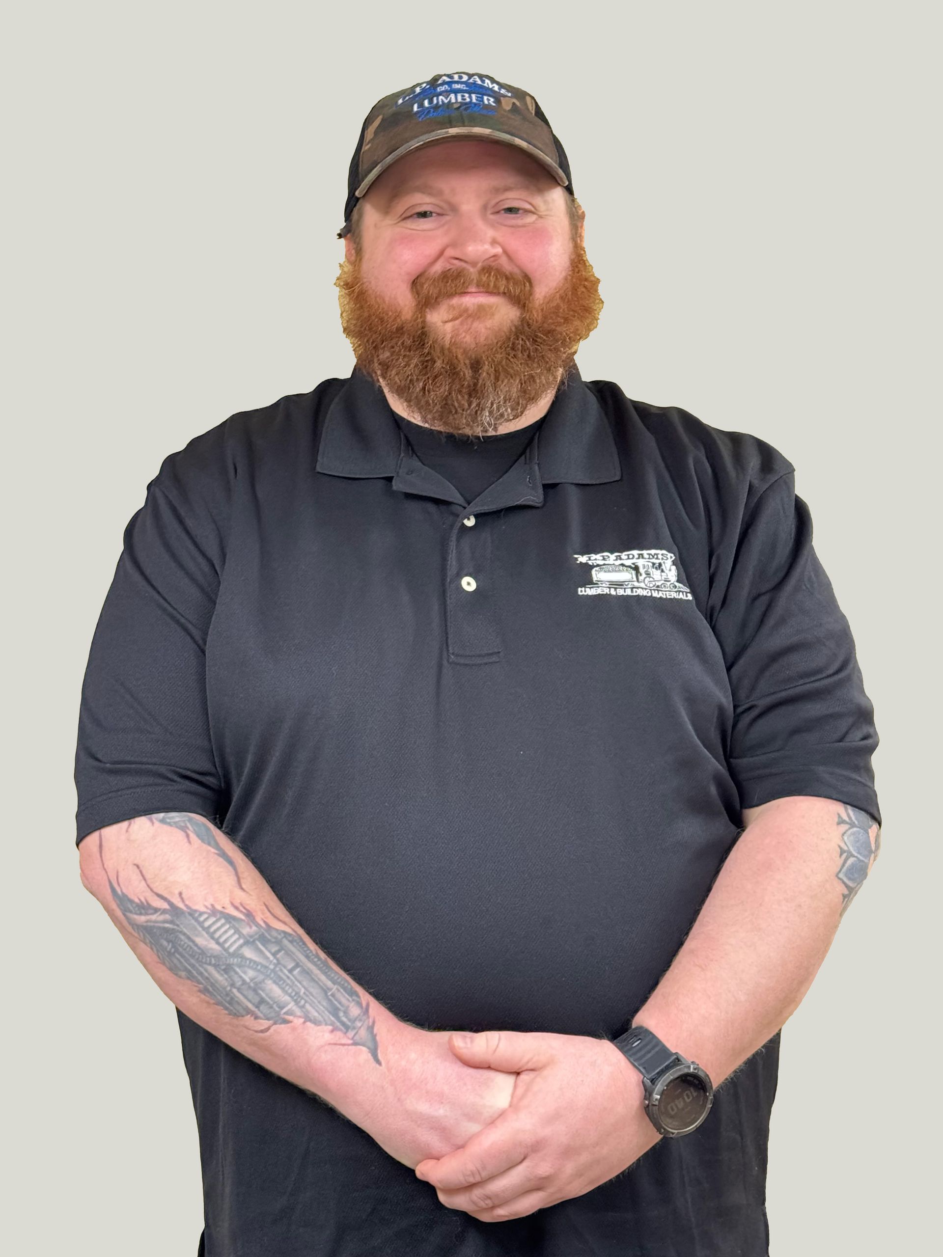 Smiling man in black polo and cap, standing with hands clasped against a plain gray background