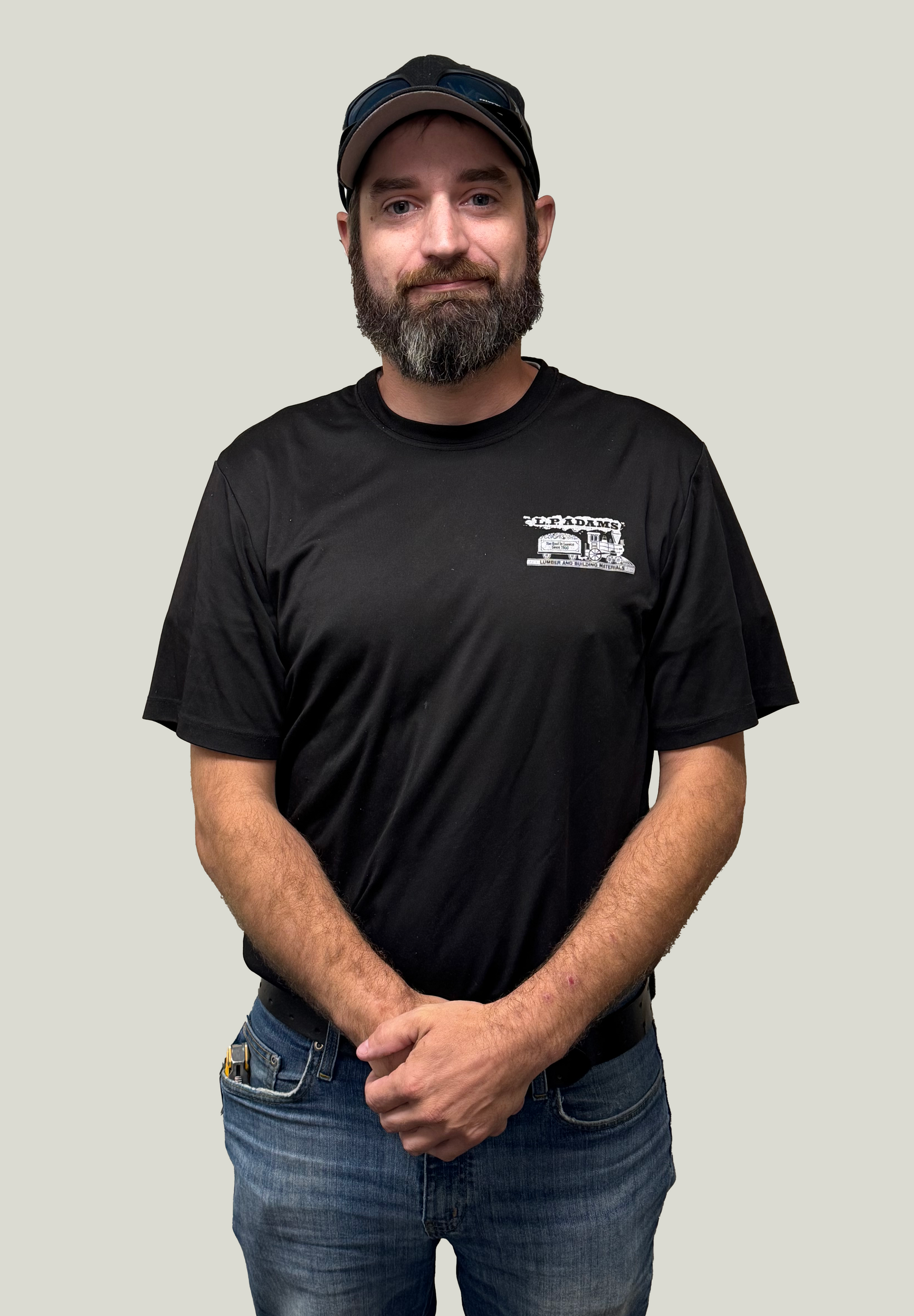 Man in black T-shirt and cap standing against a plain background with hands clasped in front