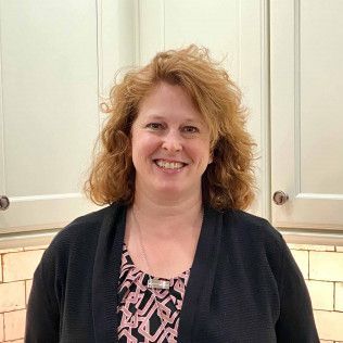 Smiling person with curly red hair standing in a kitchen, wearing a black cardigan over a patterned top.