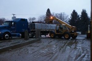 Blue truck and yellow loader unloading a flatbed truck on a muddy road in winter under gray skies