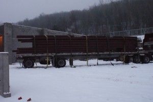 Long semi-truck parked in a snowy yard with a flatbed trailer in front of a wooded hillside