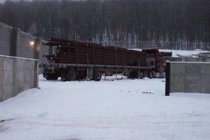 Snow-covered yard with a large truck parked beside concrete walls and a wooded hillside in the background