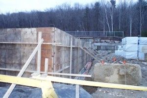 Construction site with concrete foundation walls, wooden formwork, and yellow caution tape in the foreground.
