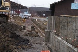 Construction site with excavator, concrete foundation, and muddy trench beside industrial buildings