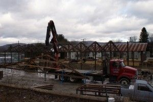 Red truck with crane lifting lumber in a muddy industrial yard under cloudy skies.