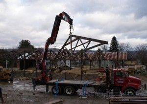 Red crane truck lifting a steel roof frame at a construction site under cloudy skies