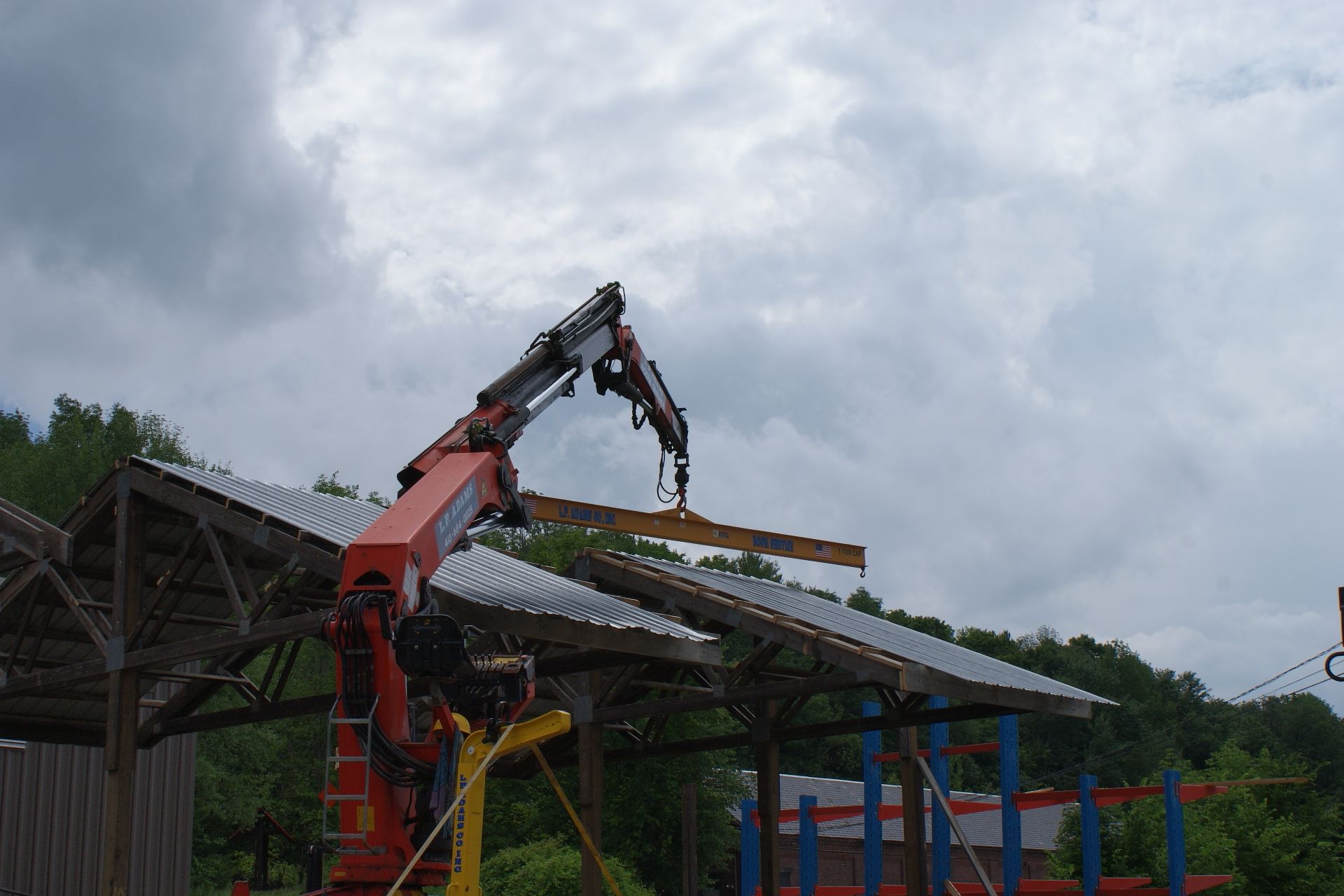 Orange crane lifting a beam onto a metal roof frame under a cloudy sky