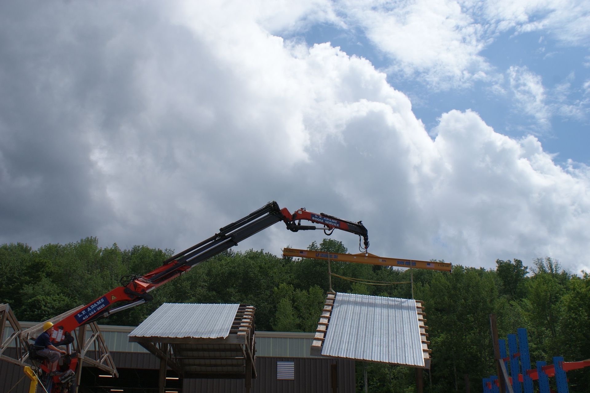 Crane lifting an orange beam over rooftops with cloudy sky and trees in the background