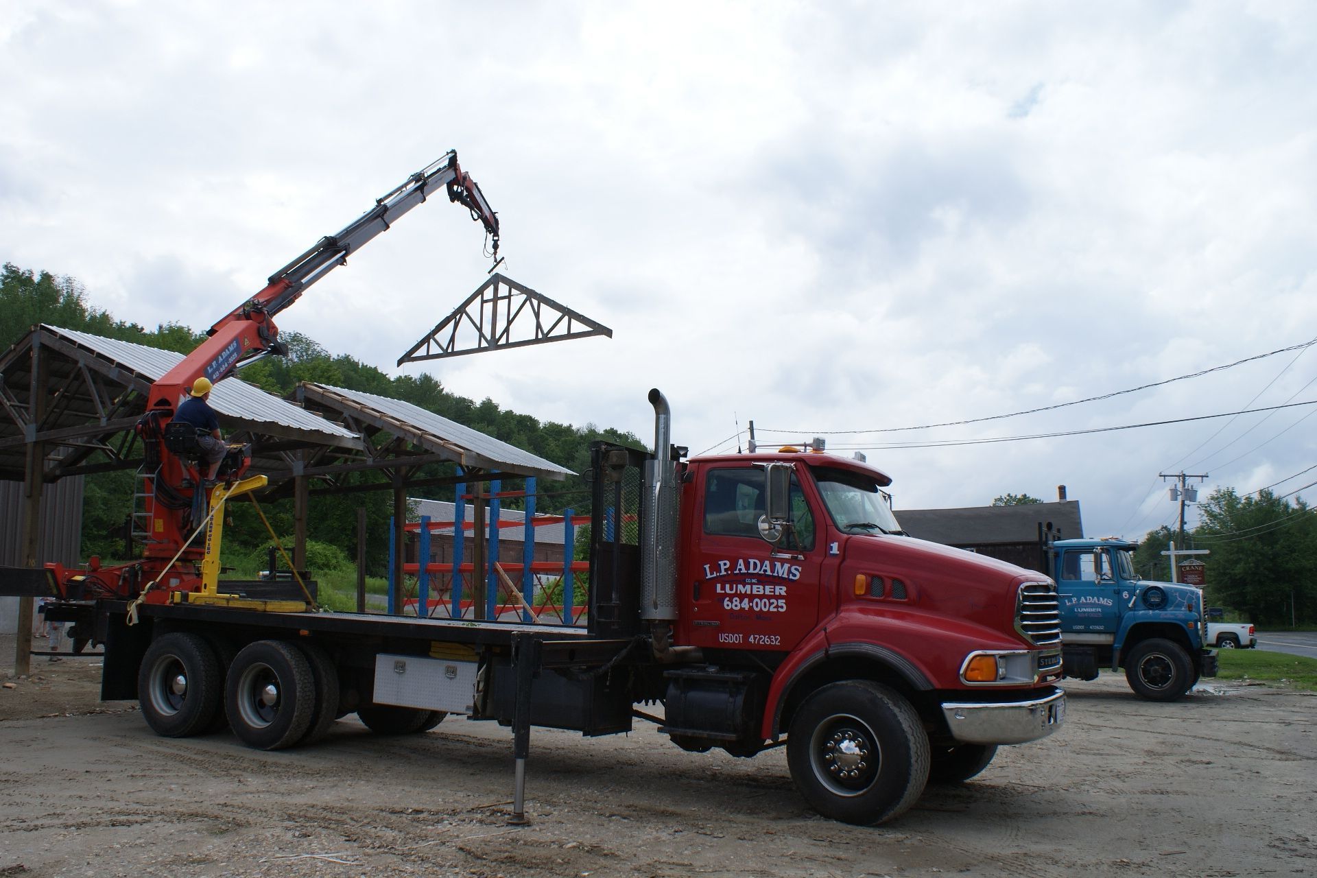 Red semi truck with crane arm carrying a load of pipes in a gravel lot under cloudy skies