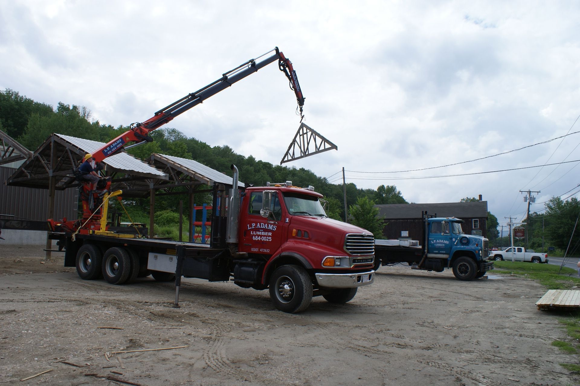 Red tow truck with crane lifting a metal frame in a gravel lot, with a blue truck in the background