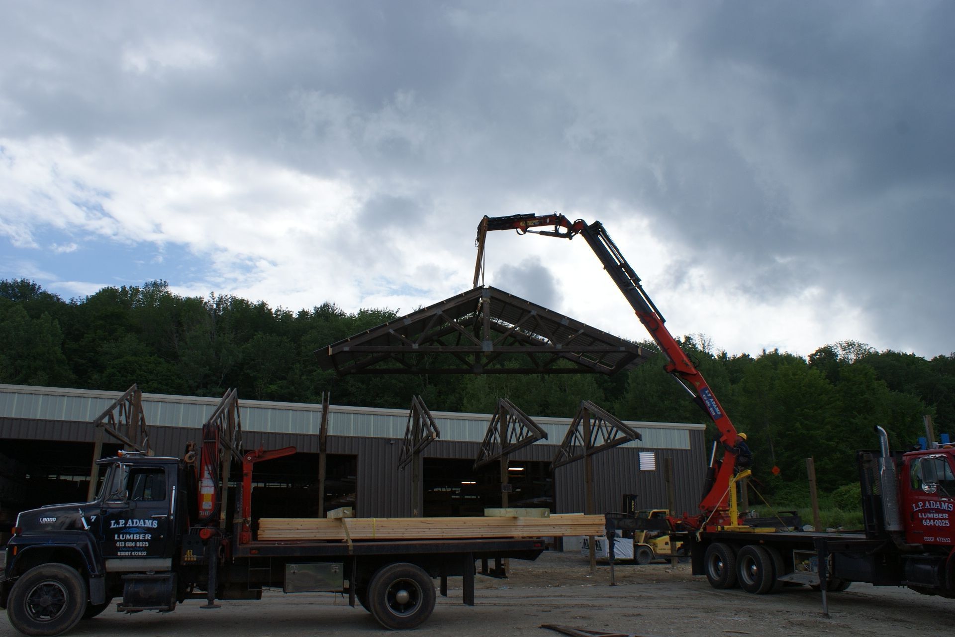 Crane lifting lumber at a construction site beside parked trucks under a cloudy sky