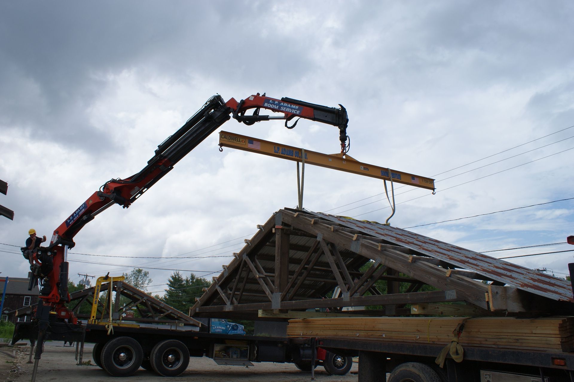 Crane truck lifting a long wooden roof truss at a construction site under a cloudy sky