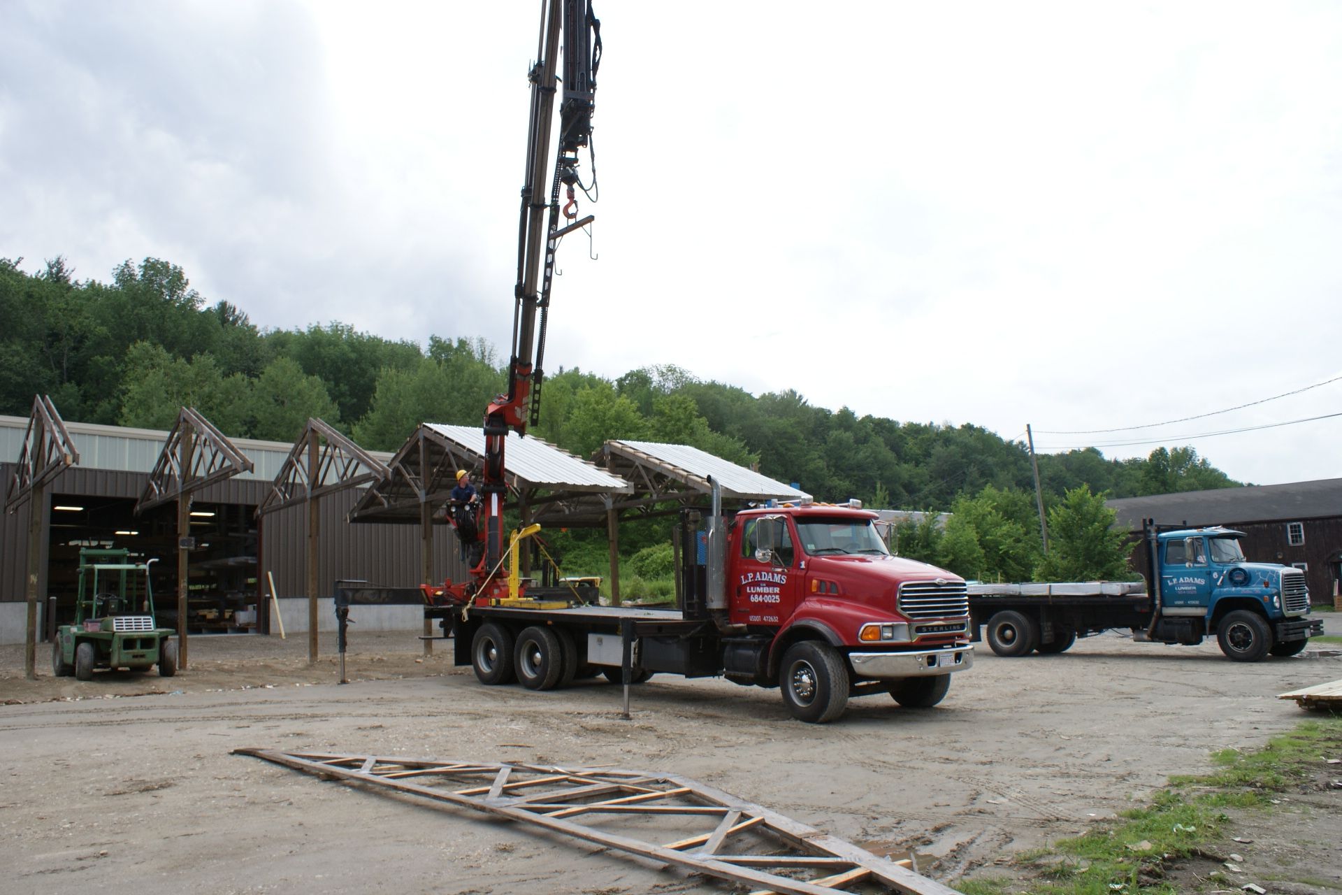 Red boom truck parked in a yard, lifting equipment near a blue truck and wooden buildings.