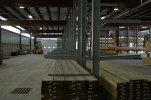 Interior of a warehouse with stacked lumber, metal beams, and palletized materials on the floor.