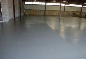 Empty warehouse interior with a smooth gray concrete floor and brick walls under skylights