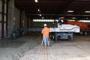 Worker in orange shirt spreading concrete inside a warehouse, with a concrete truck in the background.
