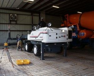 White industrial machine in a warehouse beside an orange tanker truck and tripod lights.