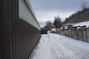 Snow-covered alley between a tall metal building and a chain-link fence, with houses in the distance