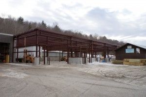 Industrial yard with a large steel-framed building under construction beside a small cabin on a snowy lot
