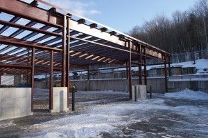 Open steel-framed building under construction in a snowy lot, with trees and a fence in the background