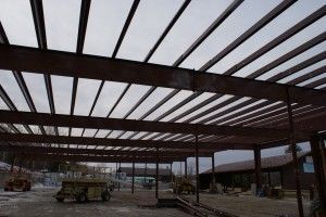 Steel building frame under construction with roof beams, columns, and scattered equipment on a muddy site