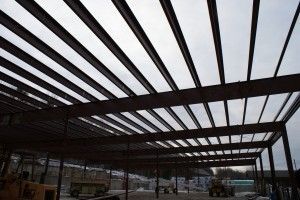 Steel frame roof under construction at a site, with beams overhead and cloudy sky visible through gaps
