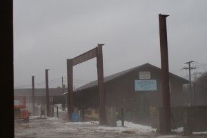 Snowy industrial yard with dark buildings and tall metal posts in the foreground