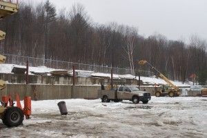 Snowy construction site with a pickup truck and excavator near a concrete wall under bare trees