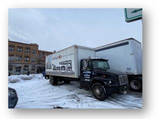 Two black delivery trucks parked in a snowy city lot beside brick buildings.