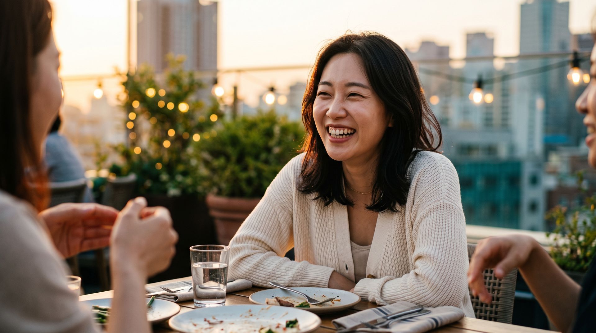 Friends laughing at an outdoor rooftop dinner at sunset, with plates, glasses, and city buildings in the background.