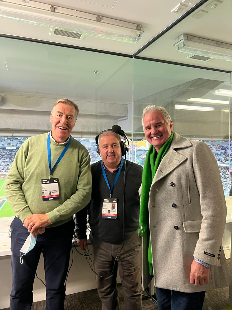 Three men in a stadium booth; two smiling, one wearing a green scarf, all with lanyards.