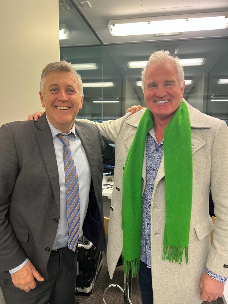 Two men smiling, one in a suit with a striped tie, the other in a coat and green scarf.
