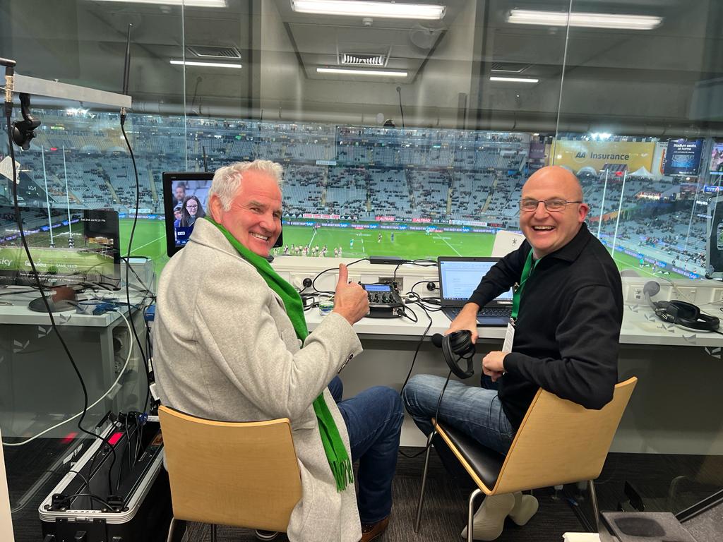 Two men in a broadcast booth overlooking a stadium. One gives a thumbs up, both smile.
