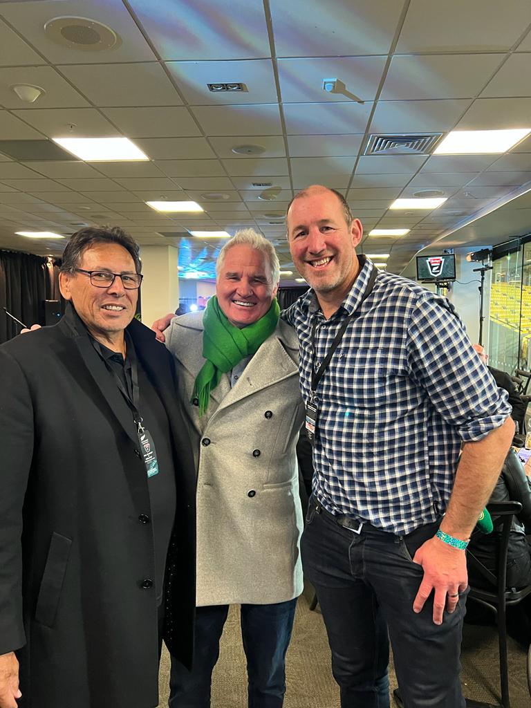 Three men in coats and scarves pose indoors, one with a green scarf and gray coat.