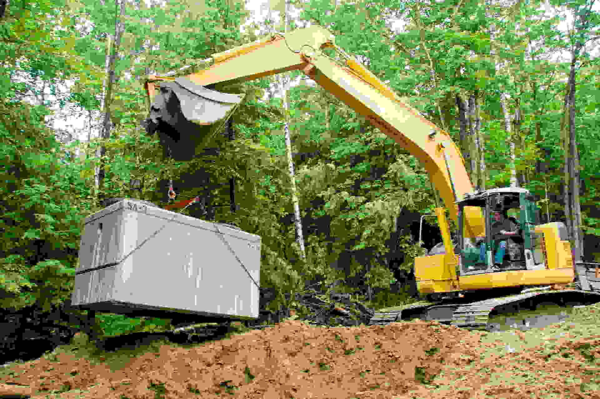 Yellow excavator lifting a gray container in a wooded construction area