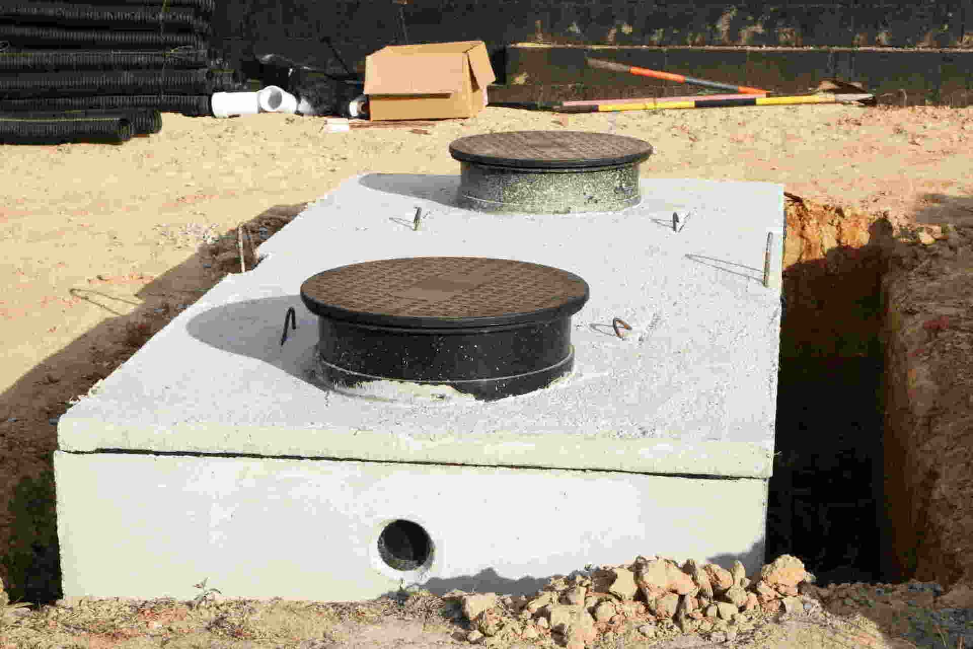 Two round manhole covers on a white concrete septic tank set in sandy ground.