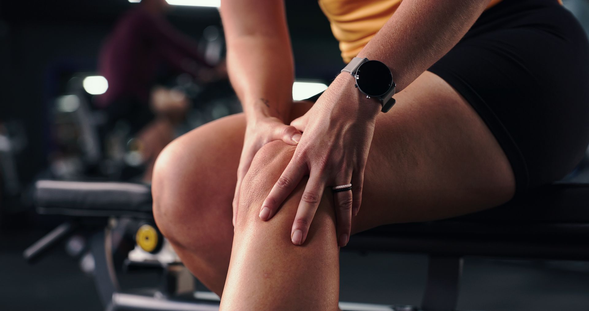 Pessoa massageando um joelho dolorido em uma academia, usando um smartwatch e uma blusa amarela.