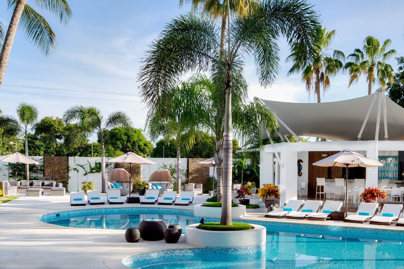 Piscina al aire libre de H2Otel by LD, rodeada de tumbonas blancas y palmeras, bajo un cielo azul claro.