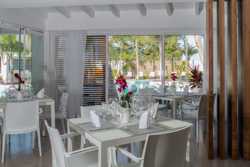 Elegante comedor interior de H2Otel, con mesas blancas y vista a palmeras, reflejando un ambiente moderno y acogedor.