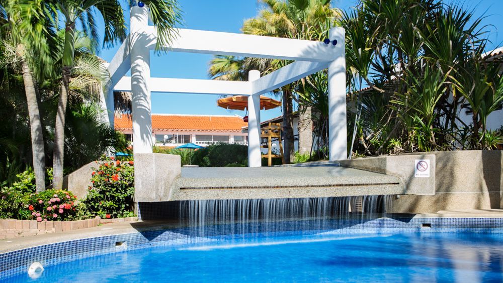 Piscina con cascada en el hotel LD Palm Beach, rodeada de vegetación tropical y ambiente relajante.